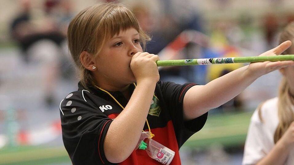 Blasrohrschießen Mädchen bläst in eine grüne Pfeife, trägt ein schwarzes Sporttrikot und eine Medaille um den Hals.