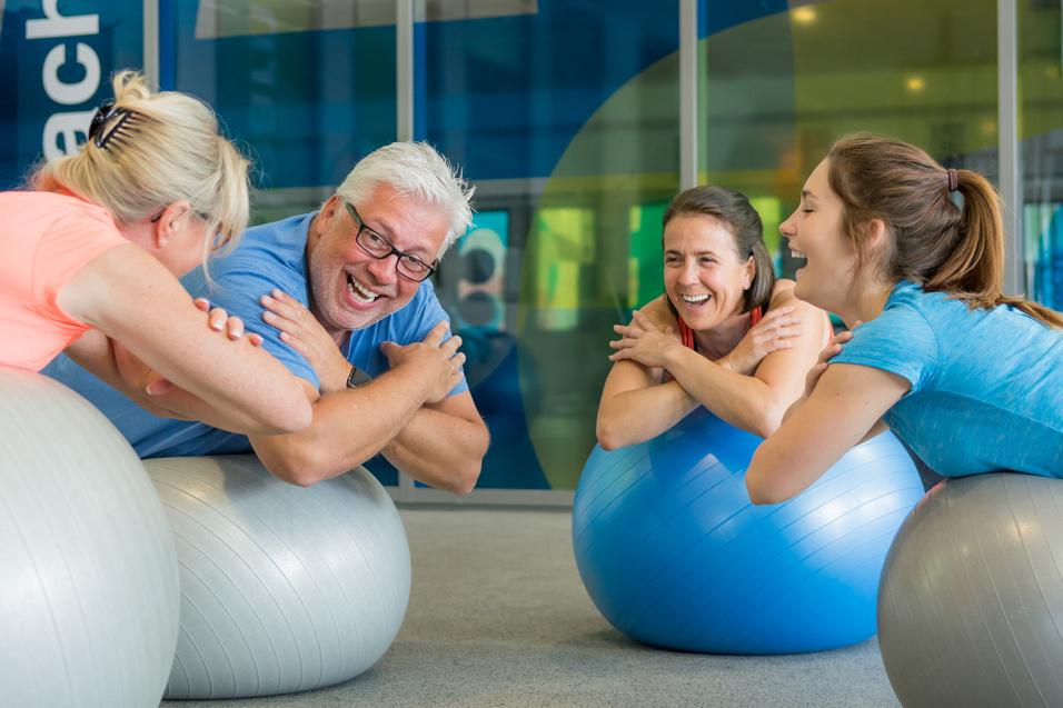 Vier Personen sitzen lachend auf Gymnastikb&auml;llen in einem hellen Fitnessraum und interagieren spielerisch miteinander.