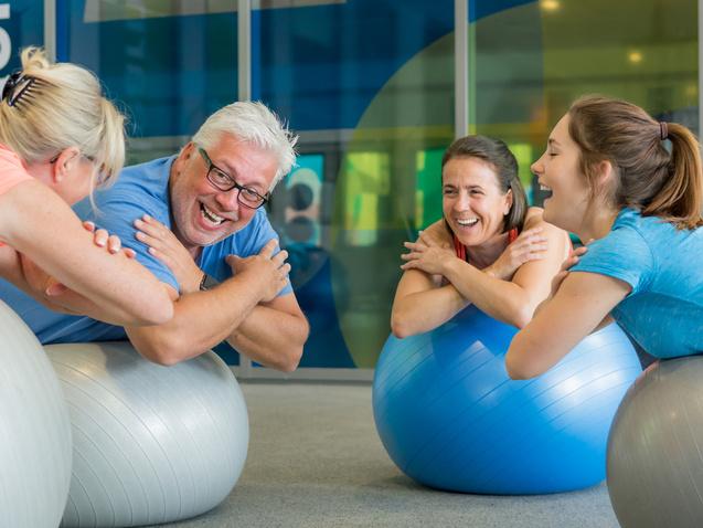 Vier Personen sitzen lachend auf Gymnastikbällen in einem hellen Fitnessraum und interagieren spielerisch miteinander.