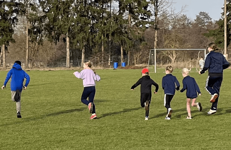 TSV Leichtathletik U8-U11 Sechs Kinder und ein Erwachsener laufen auf einem grünen Fußballfeld, umgeben von Bäumen und einem Tor im Hintergrund.