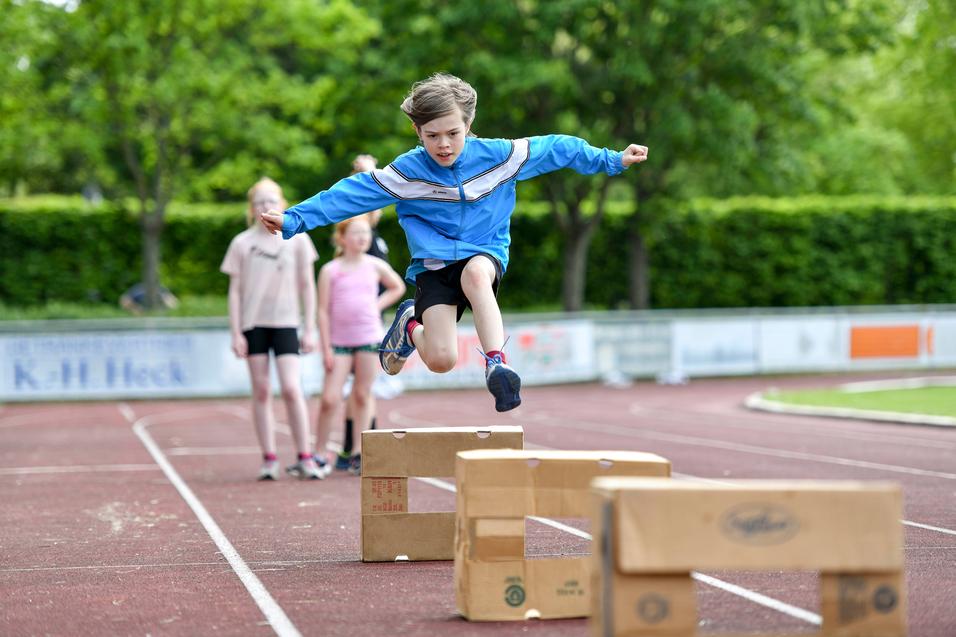 Junge springt über Holzkisten auf einer Laufbahn, während andere Kinder im Hintergrund zuschauen.