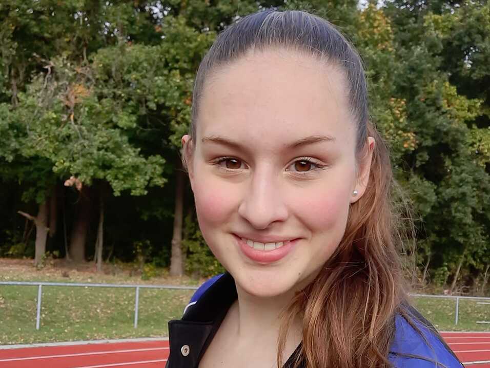 Portr&auml;t einer l&auml;chelnden jungen Frau in Sportkleidung auf einer roten Laufbahn mit B&auml;umen im Hintergrund.
