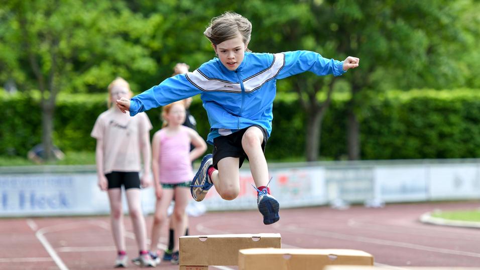 Junge springt &uuml;ber Holzkisten auf einer Laufbahn, w&auml;hrend andere Kinder im Hintergrund zuschauen.