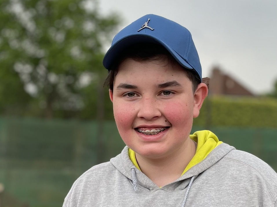 L&auml;chelnder Junge mit Kappe und grauem Sweatshirt steht an einem Tennisplatz im Freien.