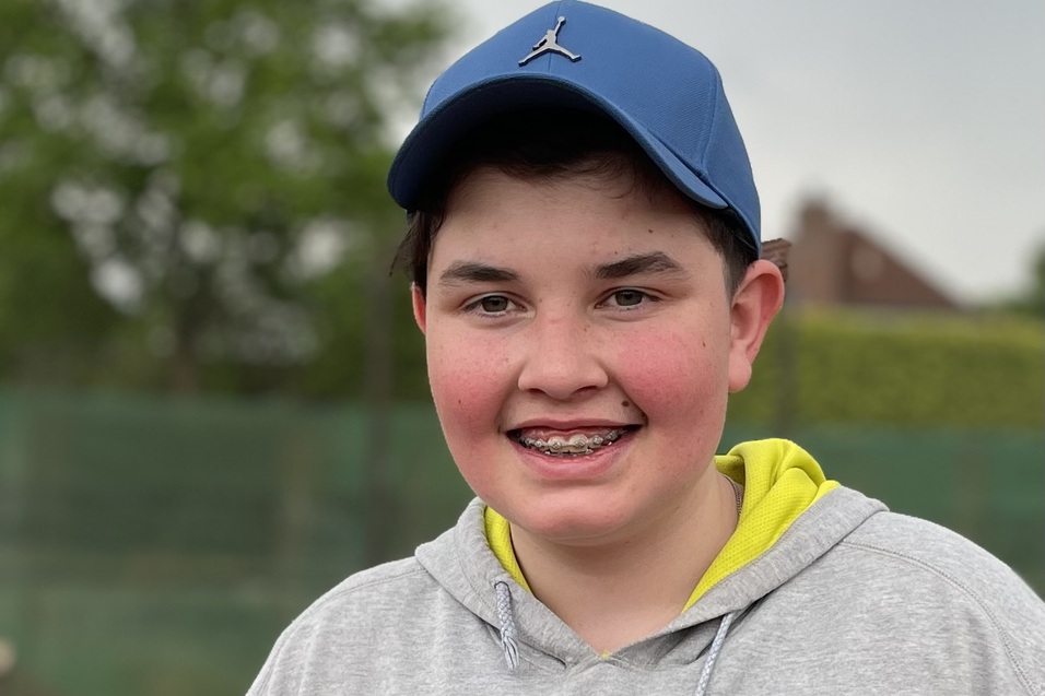 L&auml;chelnder Junge mit Kappe und grauem Sweatshirt steht an einem Tennisplatz im Freien.