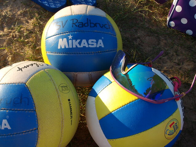 Drei Volleyballs mit unterschiedlichen Designs und einer Sonnenbrille auf sandigem Boden.