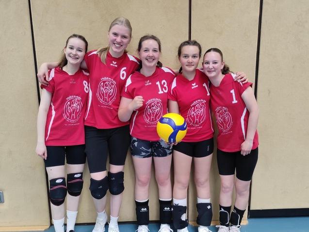 Fünf Mädchen in roten Volleyballtrikots posieren gemeinsam mit einem Volleyball in der Sporthalle.