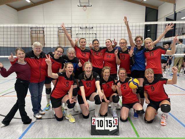 Gruppe von Volleyballspielerinnen in roten Trikots posiert jubelnd in einer Halle mit Punktetafel.