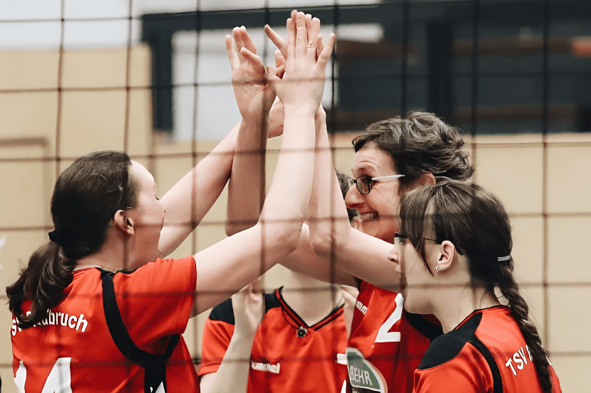 Fünf Volleyballspielerinnen in roten Trikots feiern gemeinsam an einem Netz und klatschen Hände.