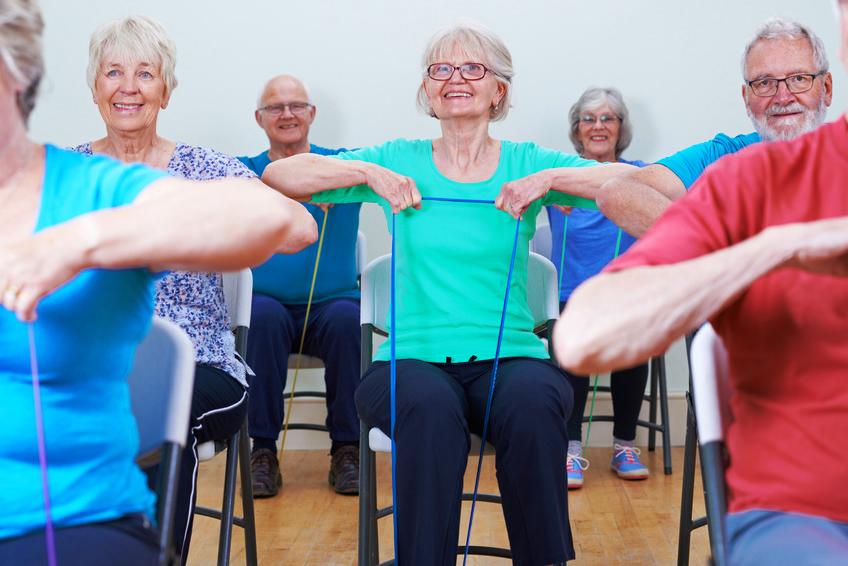 Gruppe &auml;lterer Menschen in einem Fitnesskurs, die mit einem Widerstandsband auf St&uuml;hlen sitzen und &uuml;ben.