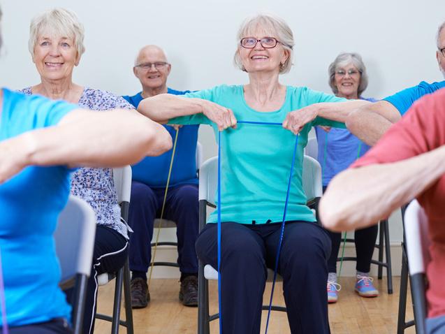 Gruppe älterer Menschen sitzt auf Stühlen und trainiert mit Widerstandsbändern in einem Fitnessraum.