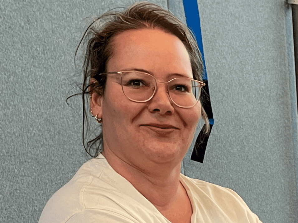 Simone Fritzsche Frau mit Brille und T-Shirt sitzt lächelnd vor einer grauen Wand, mit bunten Tattoos am Arm.