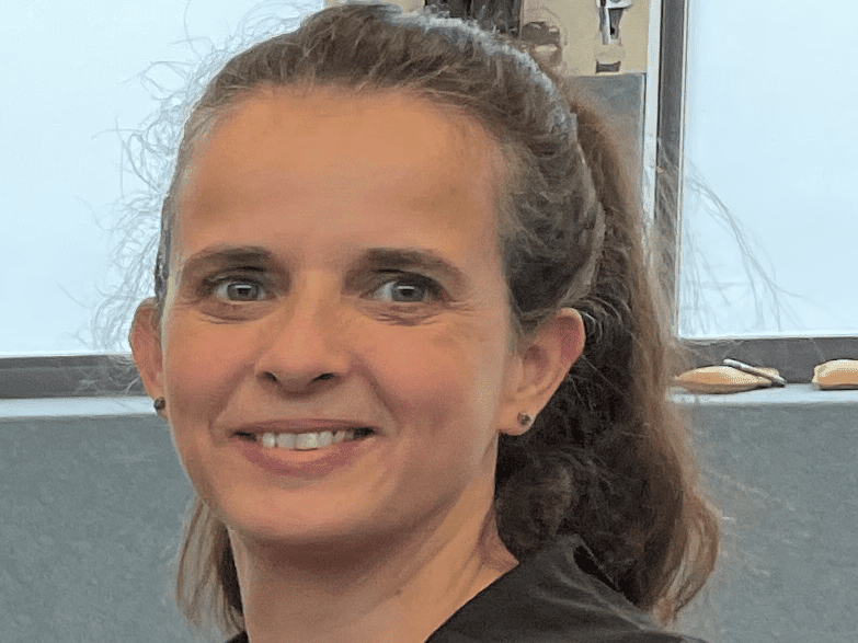 Portr&auml;t einer l&auml;chelnden Frau mit langen Haaren, die einen schwarzen Hoodie tr&auml;gt, vor einem grauen Hintergrund.