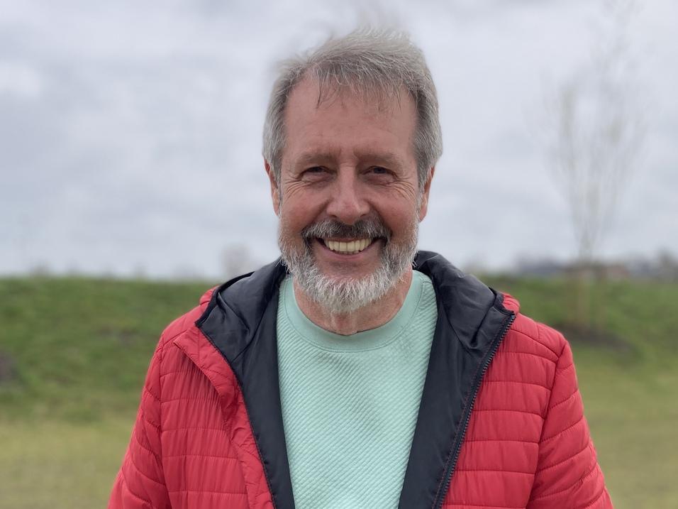Horst Karjoth Lächelnder Mann mit grauem Bart in roter Jacke und grünem Shirt vor grünem Hintergrund an einem bewölkten Tag.