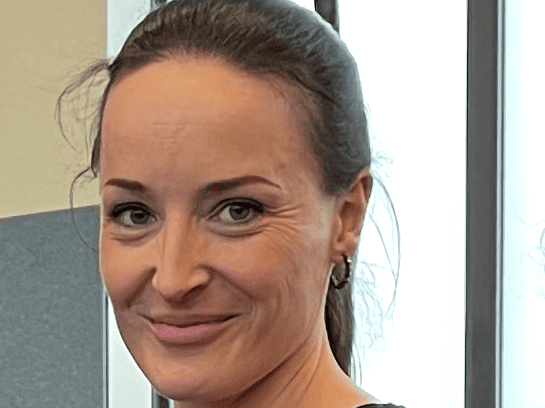Miriam Walzberg Porträt einer Frau mit langen Haaren und gestreiftem Shirt, die lächelt, vor einem Fenster in einem Raum.