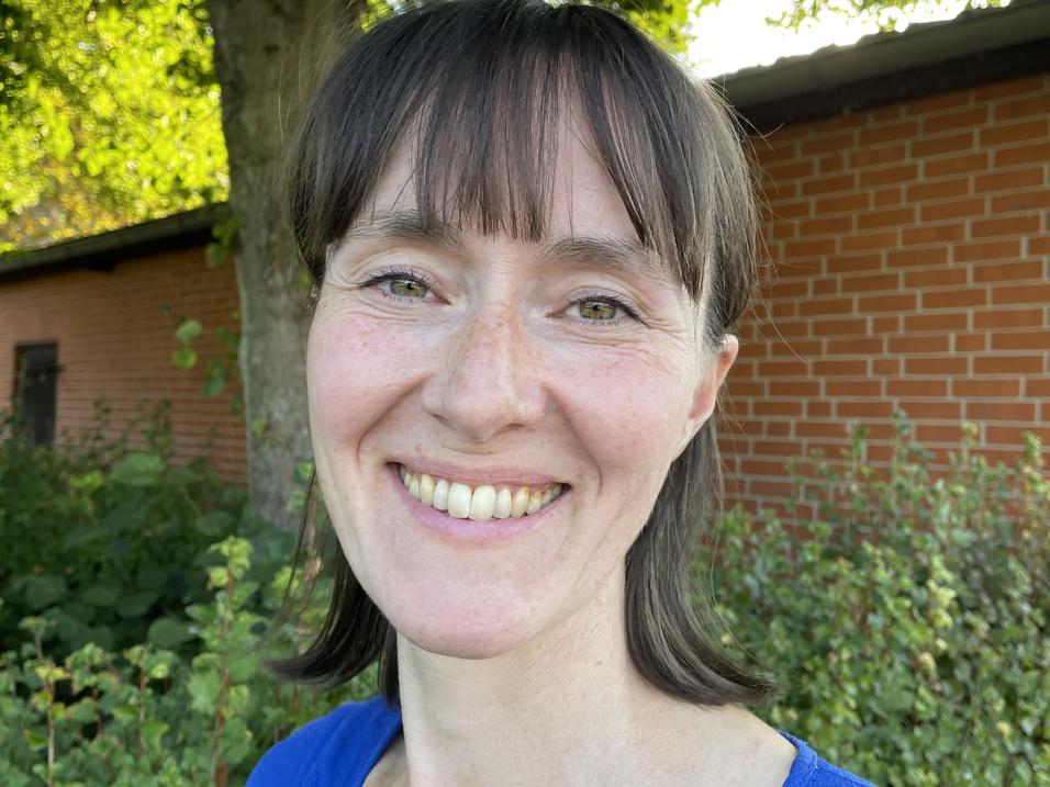 Karina Jensen Frau mit längeren Haaren und lächelndem Gesicht steht vor einem Baum und einer Backsteinmauer, umgeben von Pflanzen.