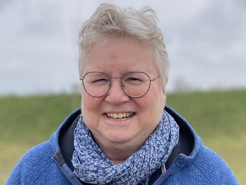 L&auml;chelnde Frau mit kurzen, grauen Haaren, tr&auml;gt einen blauen Pullover und einen Schal, vor einer gr&uuml;nen Wiese.
