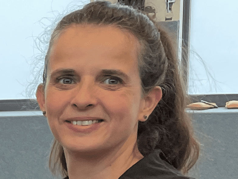 Cindy Dolke Porträt einer lächelnden Frau mit langen Haaren, die einen schwarzen Hoodie trägt, vor einem grauen Hintergrund.