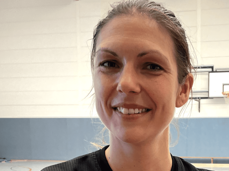 Lächelnde Frau mit sportlicher Kleidung steht in einer Sporthalle mit Basketballkorb im Hintergrund.