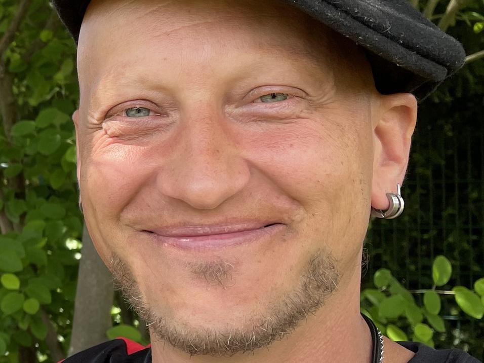 Lächelnder Mann mit Kappe und Ohrstecker vor grünem Hintergrund, in einem schwarzen Shirt mit rotem Detail.