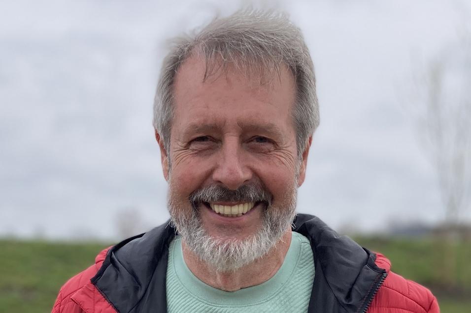 Horst Karjoth Lächelnder Mann mit grauem Bart in roter Jacke und grünem Shirt vor grünem Hintergrund an einem bewölkten Tag.