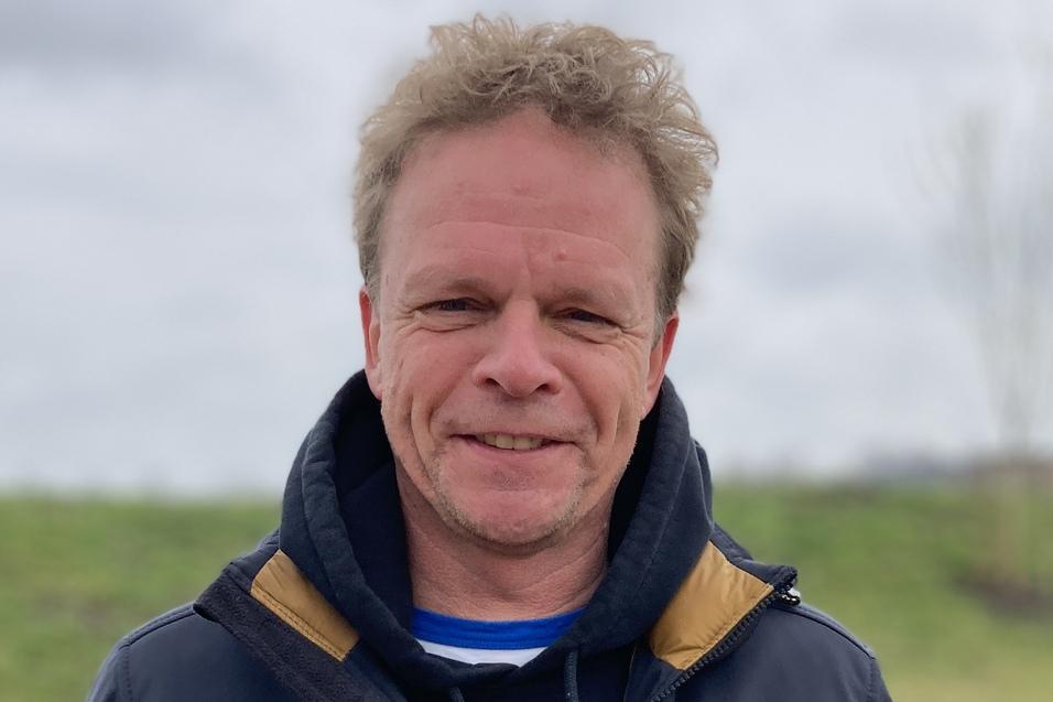 Mann mit lockigem Haar trägt eine dunkle Jacke und steht lächelnd im Freien vor einer grünen Wiese unter bewölktem Himmel.