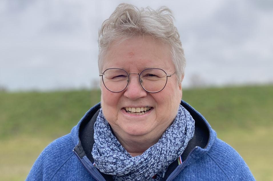 Martina Heineke Lächelnde Frau mit kurzen, grauen Haaren, trägt einen blauen Pullover und einen Schal, vor einer grünen Wiese.