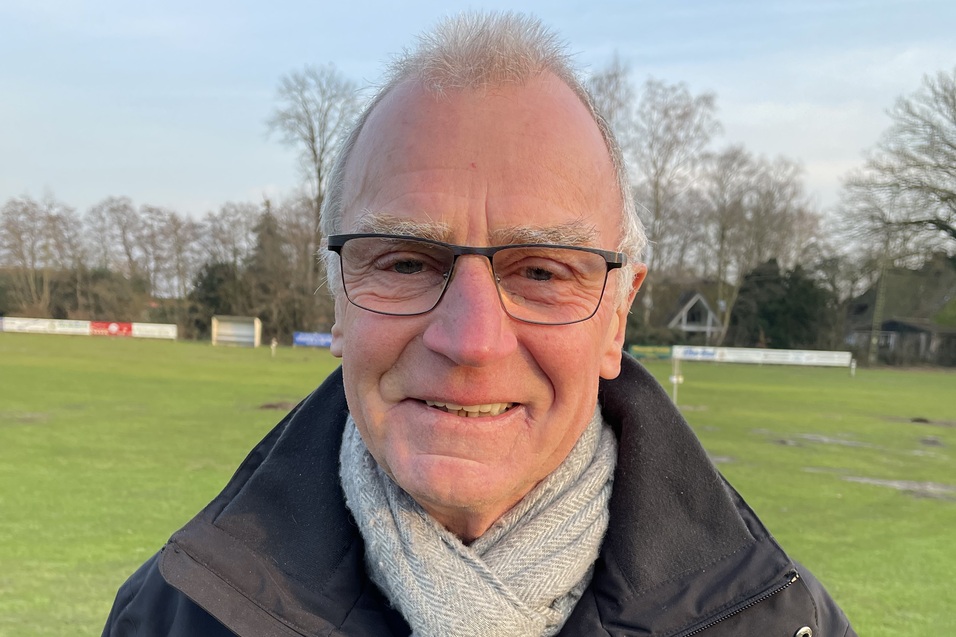L&auml;chelnder Mann mit Brille und Schal steht auf einem Fu&szlig;ballfeld, umgeben von B&auml;umen und Sporteinrichtungen im Hintergrund.