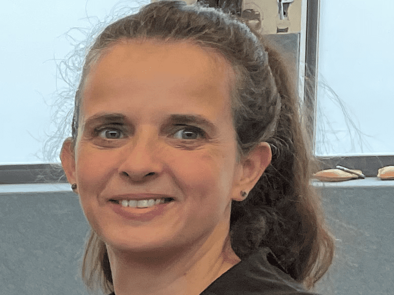 Portr&auml;t einer l&auml;chelnden Frau mit langen Haaren, die einen schwarzen Hoodie tr&auml;gt, vor einem grauen Hintergrund.