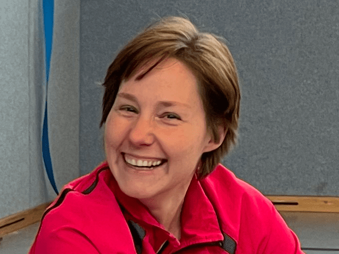 Siw Cruse Lächelnde Frau in roter Sportjacke sitzt auf dem Boden einer Sporthalle mit grauem Hintergrund.