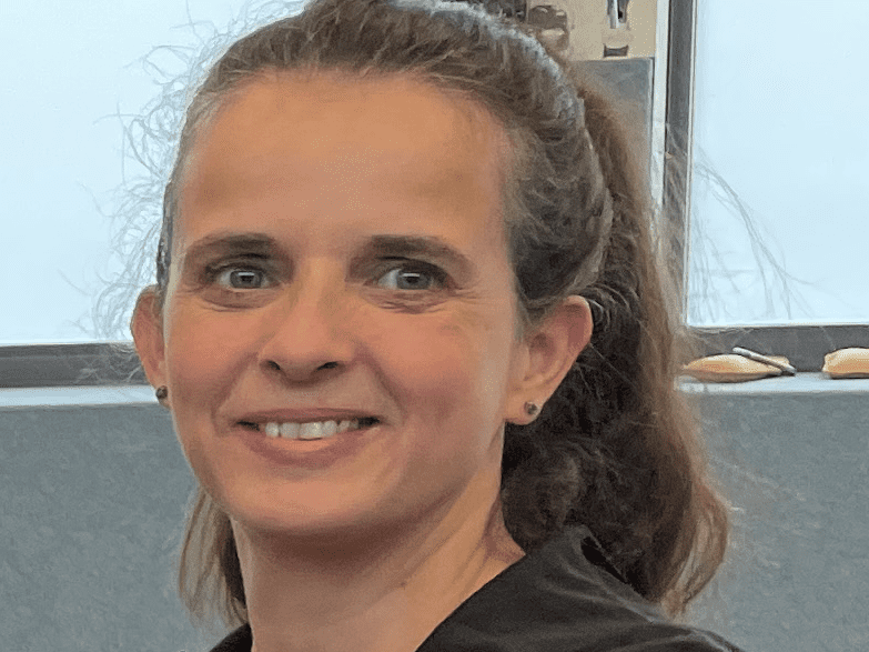 Portr&auml;t einer l&auml;chelnden Frau mit langen Haaren, die einen schwarzen Hoodie tr&auml;gt, vor einem grauen Hintergrund.