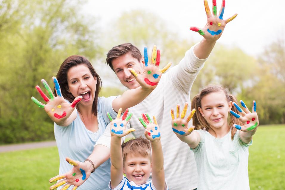 Familie mit bunten Handabdr&uuml;cken auf gr&uuml;ner Wiese, l&auml;chelnd und die H&auml;nde in die Luft haltend.