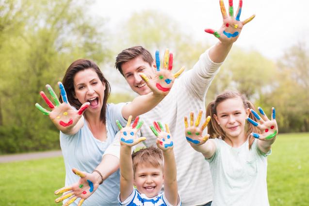 Familie mit bunten Handabdr&uuml;cken auf gr&uuml;ner Wiese, l&auml;chelnd und die H&auml;nde in die Luft haltend.