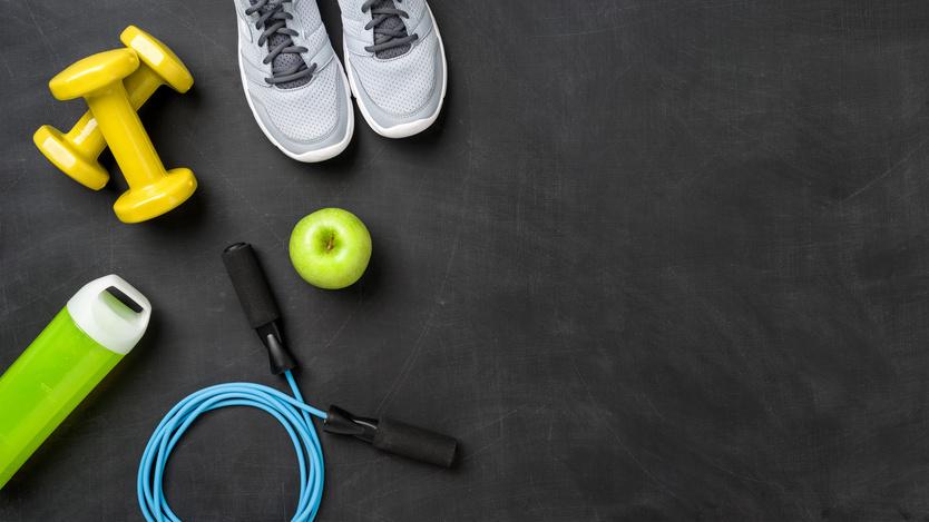 Sportzubeh&ouml;r auf einer schwarzen Unterlage: Gelbe Hantel, gr&uuml;ne Trinkflasche, blaue Springseil, Apfel und Laufschuhe.