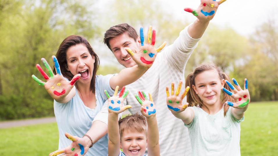 Familie mit bunten Handabdr&uuml;cken auf gr&uuml;ner Wiese, l&auml;chelnd und die H&auml;nde in die Luft haltend.