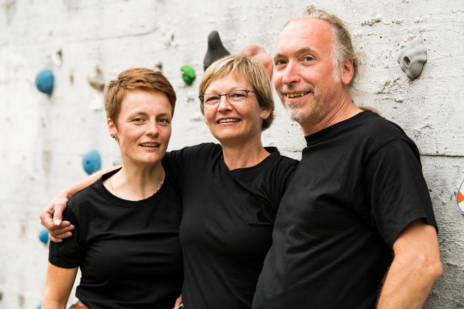 Drei Personen in schwarzen T-Shirts stehen lachend vor einer Kletterwand mit bunten Griffen.