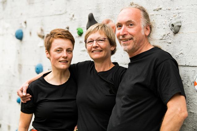 Drei Personen in schwarzen T-Shirts stehen lachend vor einer Kletterwand mit bunten Griffen.