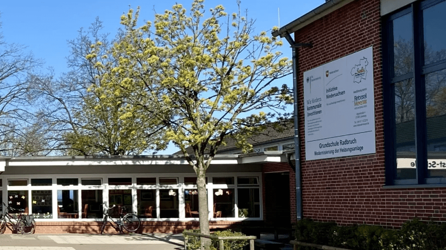 Sporthalle Grundschule Radbruch Außenansicht der Grundschule Radbruch mit Fensterfront, Baum und Fahrrad vor der Fassade an einem sonnigen Tag.