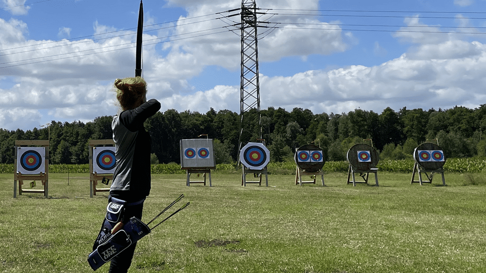 Bogenwiese Forstweg Sportliche Person mit Pfeil und Bogen in Wartestellung vor mehreren Zielscheiben auf einer Wiese.