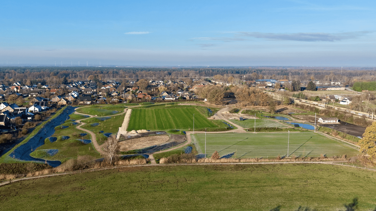 Luftaufnahme einer gr&uuml;nen Sportanlage umgeben von Wohngebieten und W&auml;ldern bei klarem Wetter.