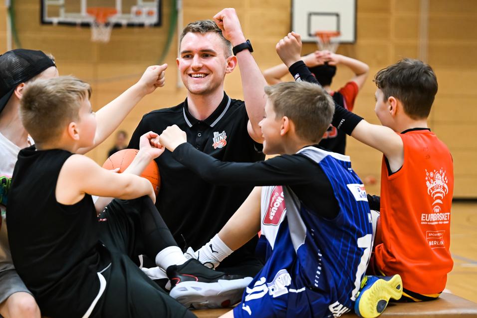 Sechs Kinder und ein Trainer zeigen Freude beim Basketballtraining auf einer Holzbank in einer Sporthalle.