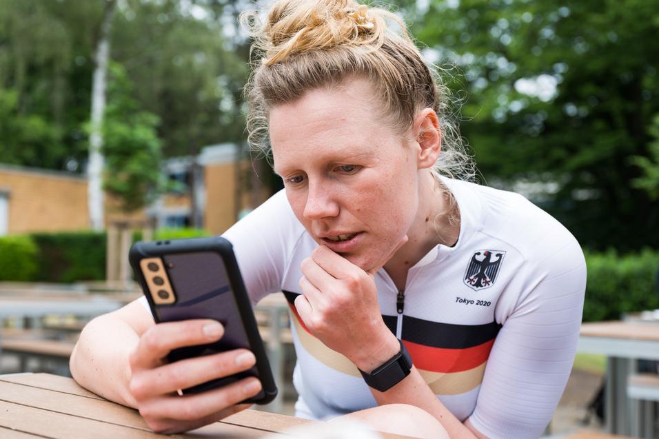 Athletin im weißen Trikot mit schwarzem und rotem Streifen schaut konzentriert auf ihr Smartphone in einer Außenanlage.