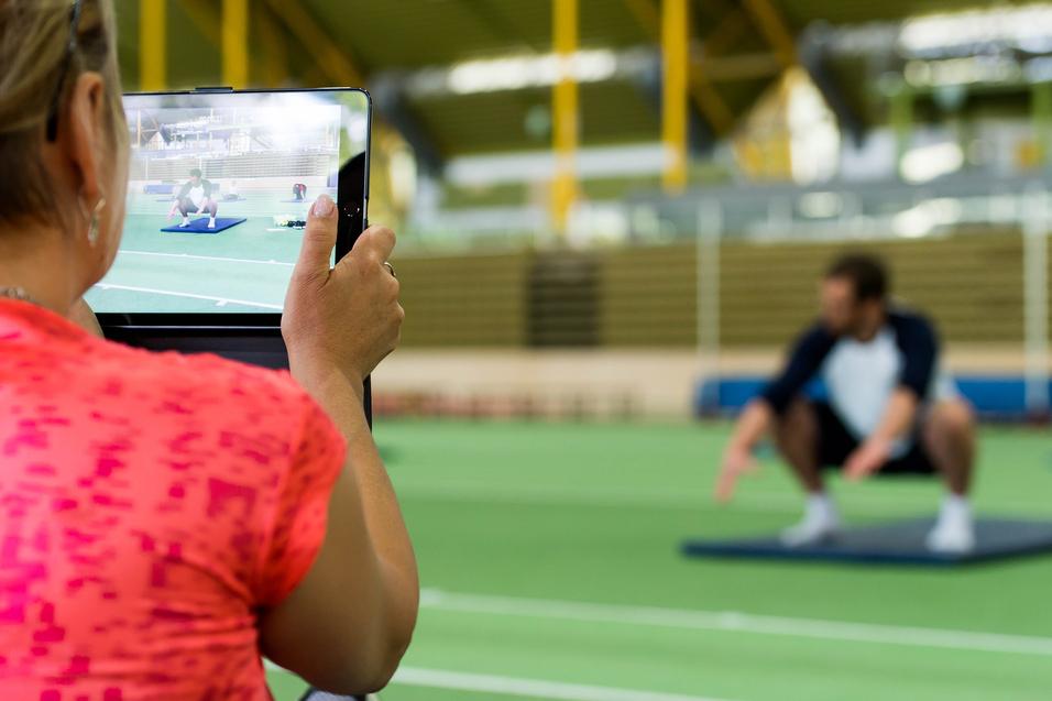 Eine Person filmt einen Mann, der auf einer Matte in der Squat-Position trainiert, in einem Sportzentrum.
