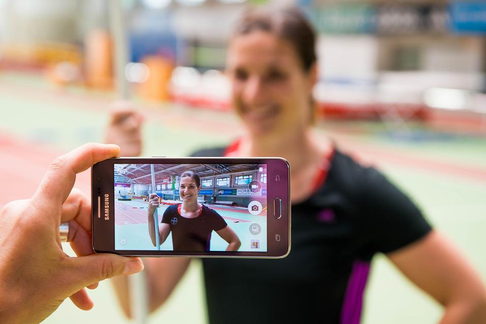 Hand hält ein Smartphone, das ein Porträt einer lächelnden Athletin mit Stab in einer Sporthalle zeigt.