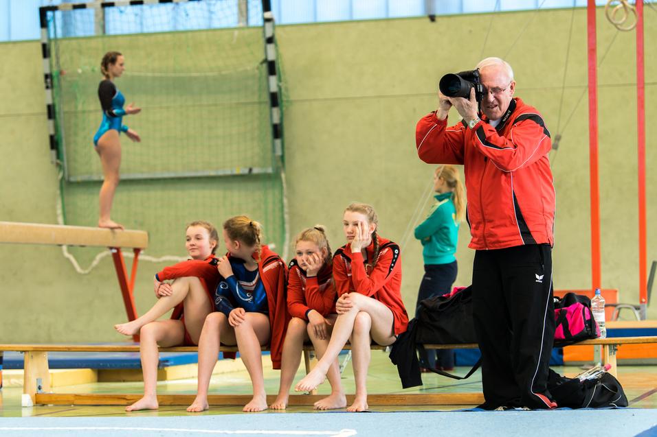 Trainer fotografiert während einer Turneinheit vier junge Turnerinnen auf einer Bank in einer Sporthalle.