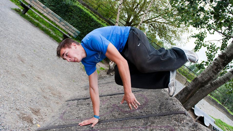 Junger Mann führt eine Parkour-Bewegung auf einer Mauer in einem Park aus, mit Bäumen und einer Bank im Hintergrund.