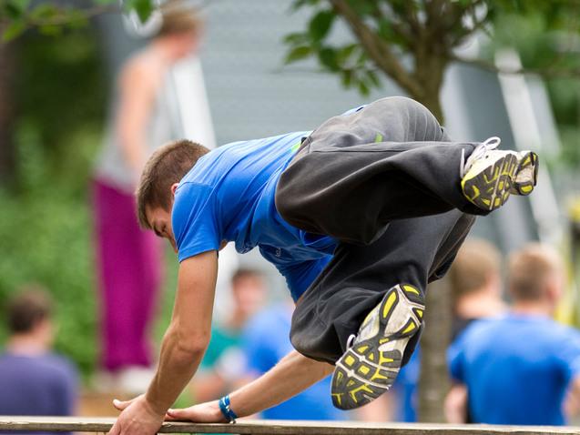 Junge in blauer T-Shirt springt über eine Holzplattform, umgeben von Bäumen und weiteren Parkour-Aktivisten.