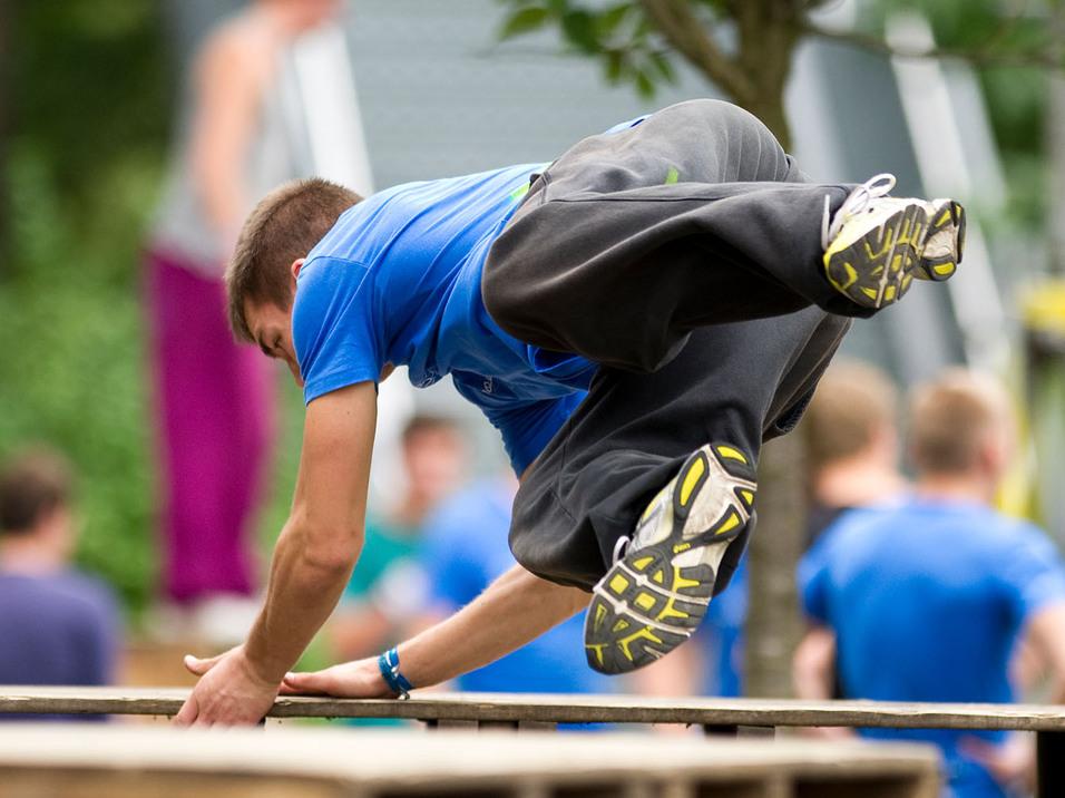 Junge in blauer T-Shirt springt über eine Holzplattform, umgeben von Bäumen und weiteren Parkour-Aktivisten.