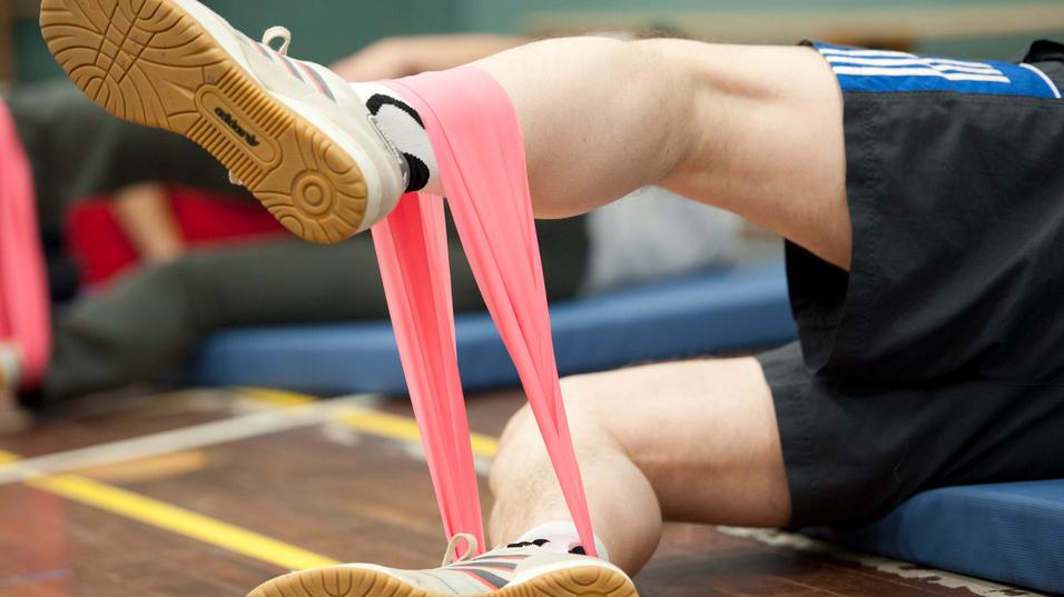 Bein eines Sportlers mit pinkem Widerstandsband, auf einer Sportmatte, im Hintergrund weitere Teilnehmer.