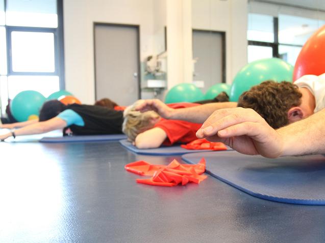 Gruppentraining mit Personen liegend auf Matten, während Bälle im Hintergrund sichtbar sind.